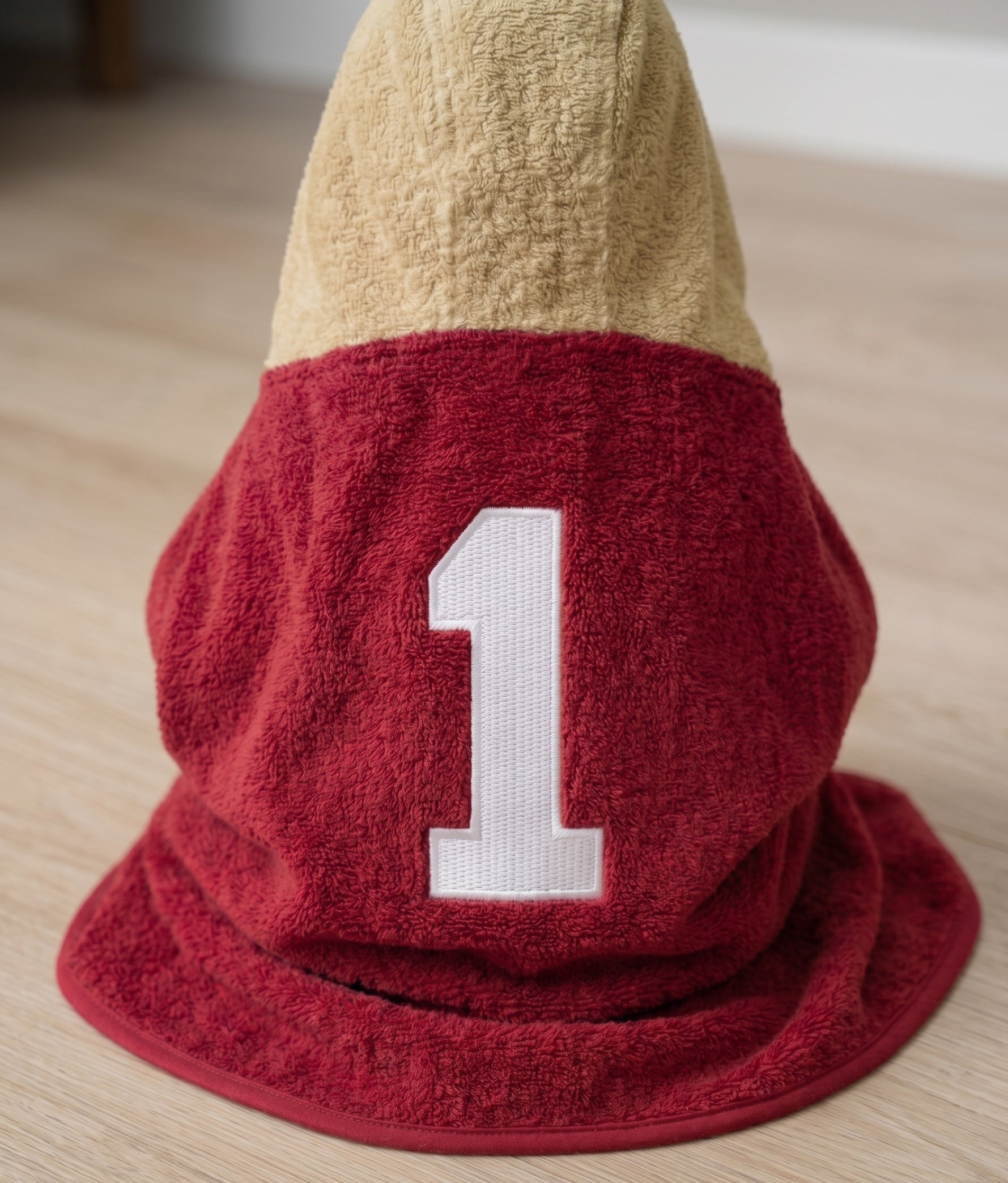 Baby wearing a hooded towel withgarnet lower section, and a large white embroidered number 1, photographed on a light wood floor