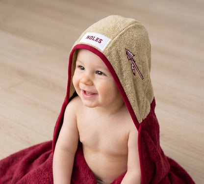A smiling baby wrapped in a garnet towel and wearing a beige hooded towel with a white patch reading 'NOLES' and a spear  logo, seated on a light wood floor.