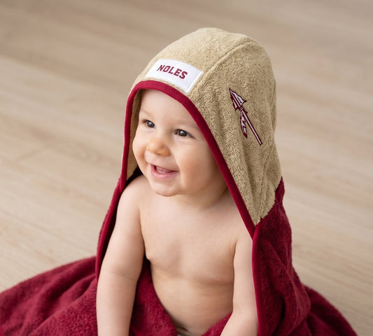 A smiling baby wrapped in a garnet towel and wearing a beige hooded towel with a white patch reading 'NOLES' and a spear  logo, seated on a light wood floor.